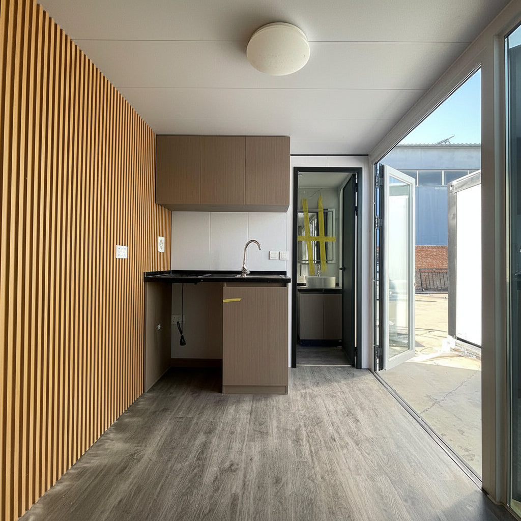 A compact kitchenette inside a mobile home or modular unit, featuring light brown wood cabinets, a black countertop with a sink, gray wood-look flooring, and a slatted wood accent wall. Large glass doors on the right open to an outdoor patio area.