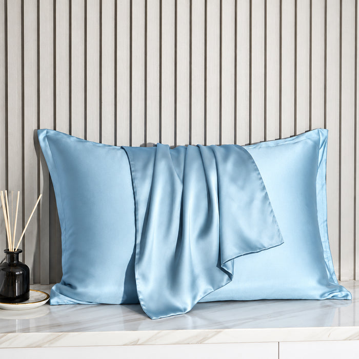 A light blue satin pillowcase with matching pillow and folded sheet, styled on a marble surface against a gray slatted wood wall.