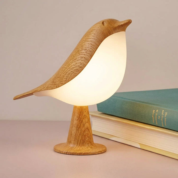 A bird-shaped table lamp made of light wood and glowing white LED, shown lit on a tabletop next to stacked books.